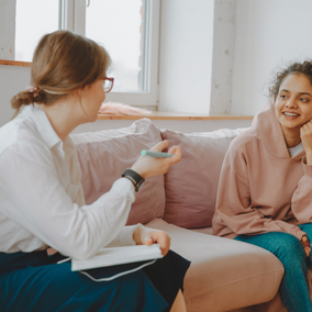 Two women sit on a pink couch, talking and smiling in a bright room.