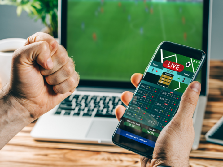Man holds smartphone showing live sports betting, clenched fist raised, laptop streams soccer game.