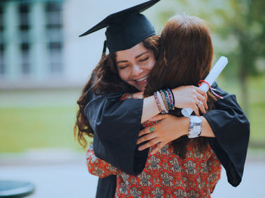 Graduate in cap and gown joyfully hugs a woman while holding a diploma, with greenery in the background.